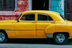 A classic yellow vintage car is parked beside a colorful and vibrant wall in a street setting, capturing the charm and nostalgia of a bygone era.