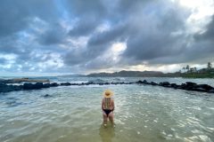 A woman in a black swimsuit and straw hat stands in shallow ocean water facing away under a dramatic cloudy sky, capturing a moment of reflection and connection with nature.